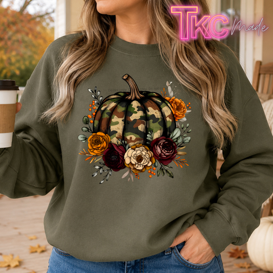 A woman wearing an olive green crewneck sweatshirt with a large autumn graphic of a camouflage pumpkin surrounded by orange, burgundy, and cream fall flowers. She is holding a coffee cup in a porch setting.