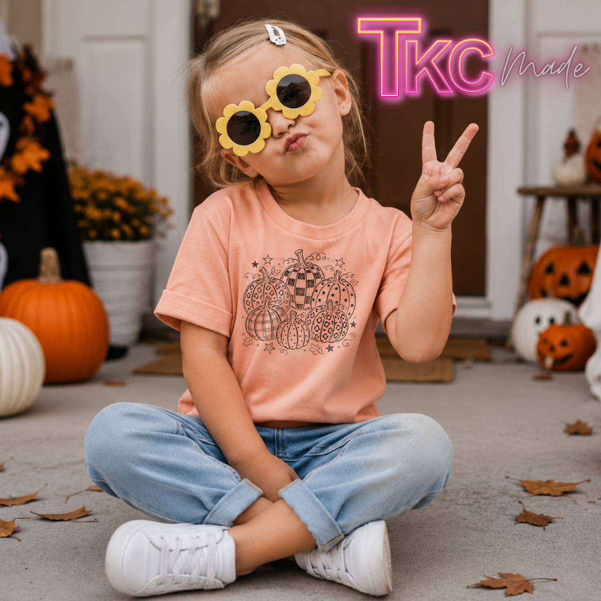 A young child sitting cross-legged outdoors wearing a peach graphic T-shirt, light blue jeans, and white sneakers, with yellow flower-shaped sunglasses. Pumpkins and autumn decorations are visible in the background, and a pink 'TKC Made' logo appears in the top right.