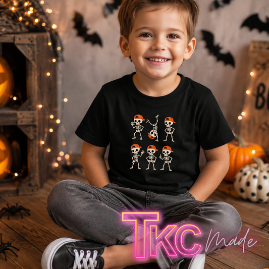 A young boy wearing a black graphic t-shirt with cartoon skeleton illustrations, sitting in a cozy Halloween-themed room with pumpkins, string lights, and bats.