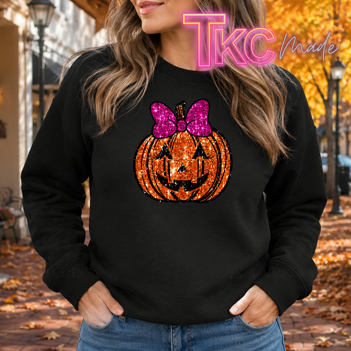 A woman wearing a black sweatshirt with a glittery orange jack-o'-lantern graphic and a pink bow on the front, standing outdoors on a street with autumn leaves and trees in the background. A 'TKC Made' watermark is visible at the top.