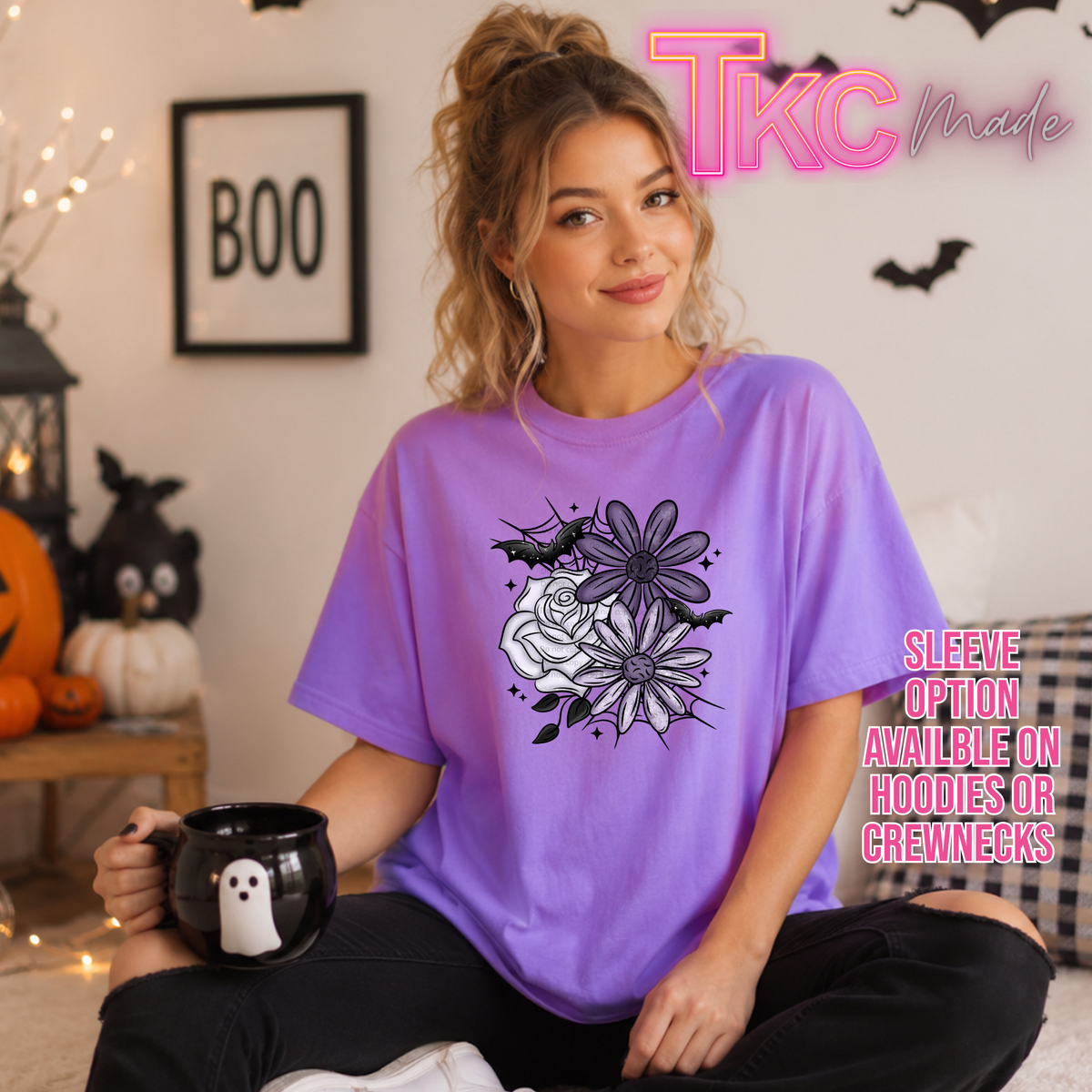 A woman wearing a purple oversized graphic T-shirt with a black and white floral design and bats, sitting in a cozy Halloween-themed room and holding a black mug with a white ghost. The shirt features a pink 'TKC Made' logo overlay in the top right, and text on the right reads 'Sleeve option available on hoodies or crewnecks'.
