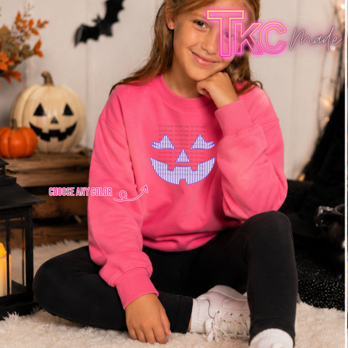 A young girl sitting on a white rug wearing a bright pink crewneck sweatshirt with a blue and white pixelated jack-o'-lantern graphic on the front. The background features Halloween decorations including a jack-o'-lantern, a lantern, and a black bat silhouette.