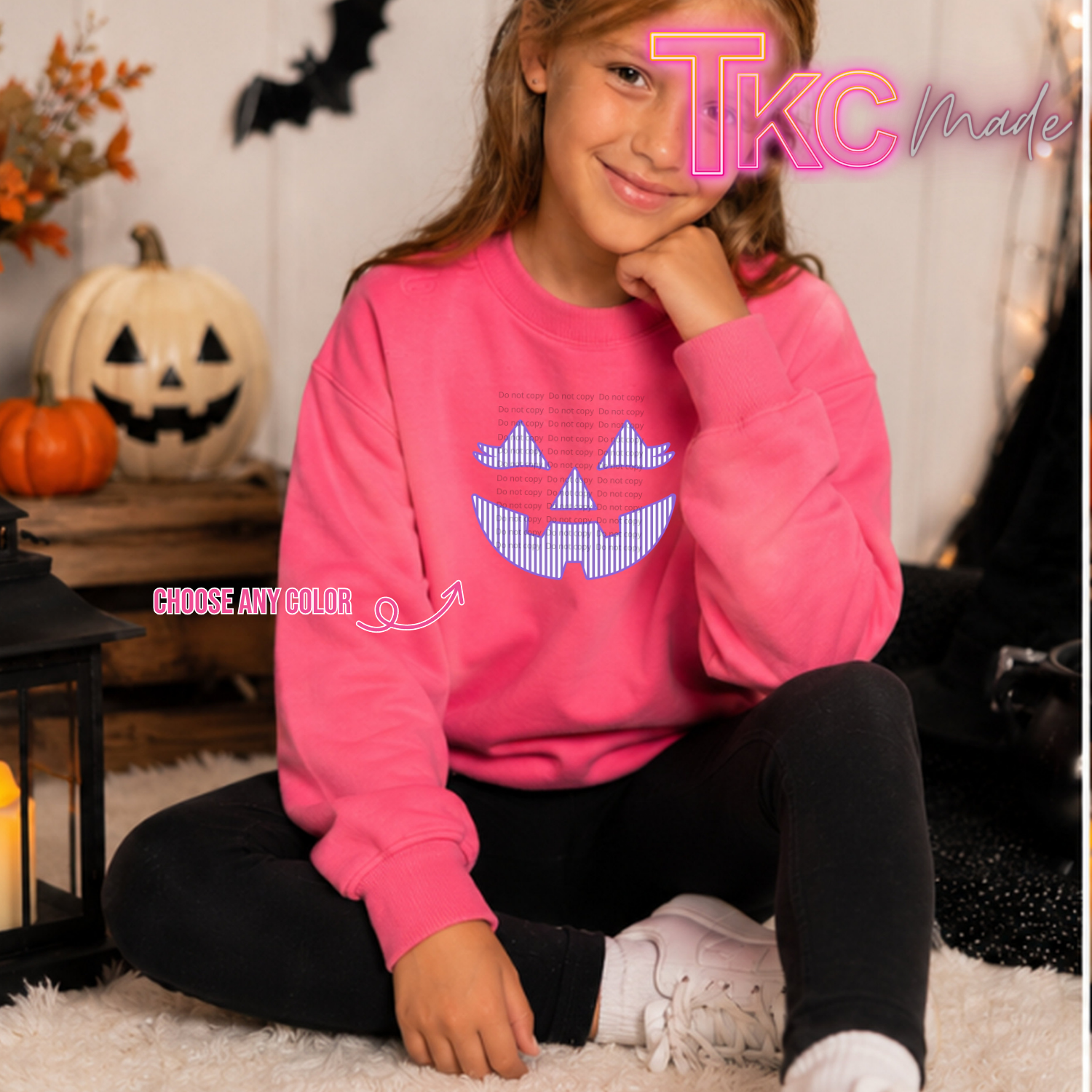 A young girl sitting on a white rug wearing a bright pink crewneck sweatshirt with a blue and white pixelated jack-o'-lantern graphic on the front. The background features Halloween decorations including a jack-o'-lantern, a lantern, and a black bat silhouette.