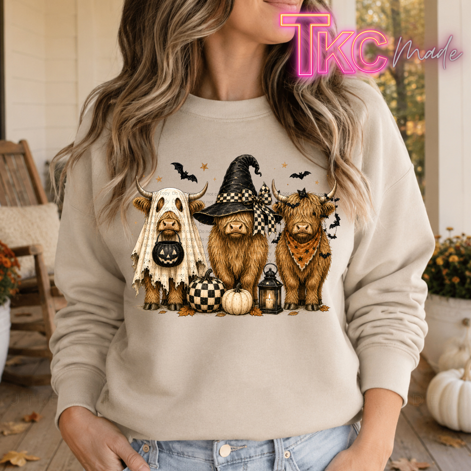 A woman wearing a beige crewneck sweatshirt with a graphic of three cute Highland cows dressed in Halloween costumes (ghost, witch hat, and bandana) with pumpkins and bats. The photo is taken on a rustic wooden porch with a 'TKC Made' watermark in the top right corner.