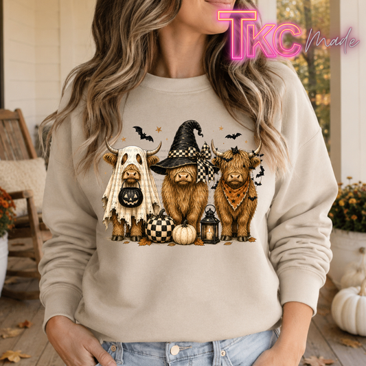 A woman wearing a beige crewneck sweatshirt with a graphic of three cute Highland cows dressed in Halloween costumes (ghost, witch hat, and bandana) with pumpkins and bats. The photo is taken on a rustic wooden porch with a 'TKC Made' watermark in the top right corner.