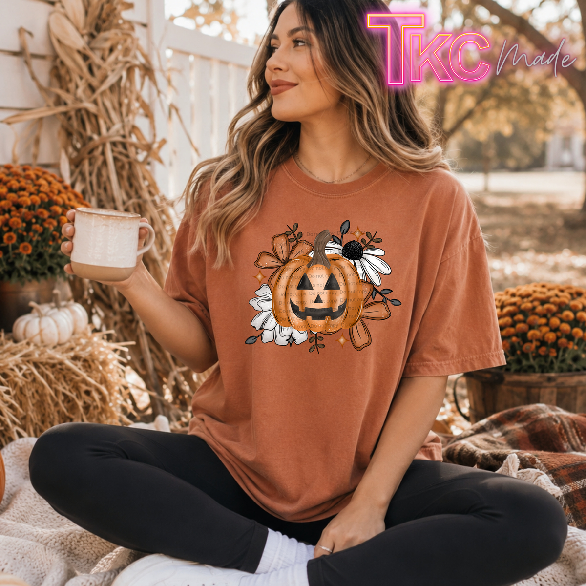 A woman sits cross-legged outdoors wearing a rust-orange graphic T-shirt with a jack-o'-lantern and autumn floral design, holding a mug. The background shows fall decorations including mums, straw, and a white fence.
