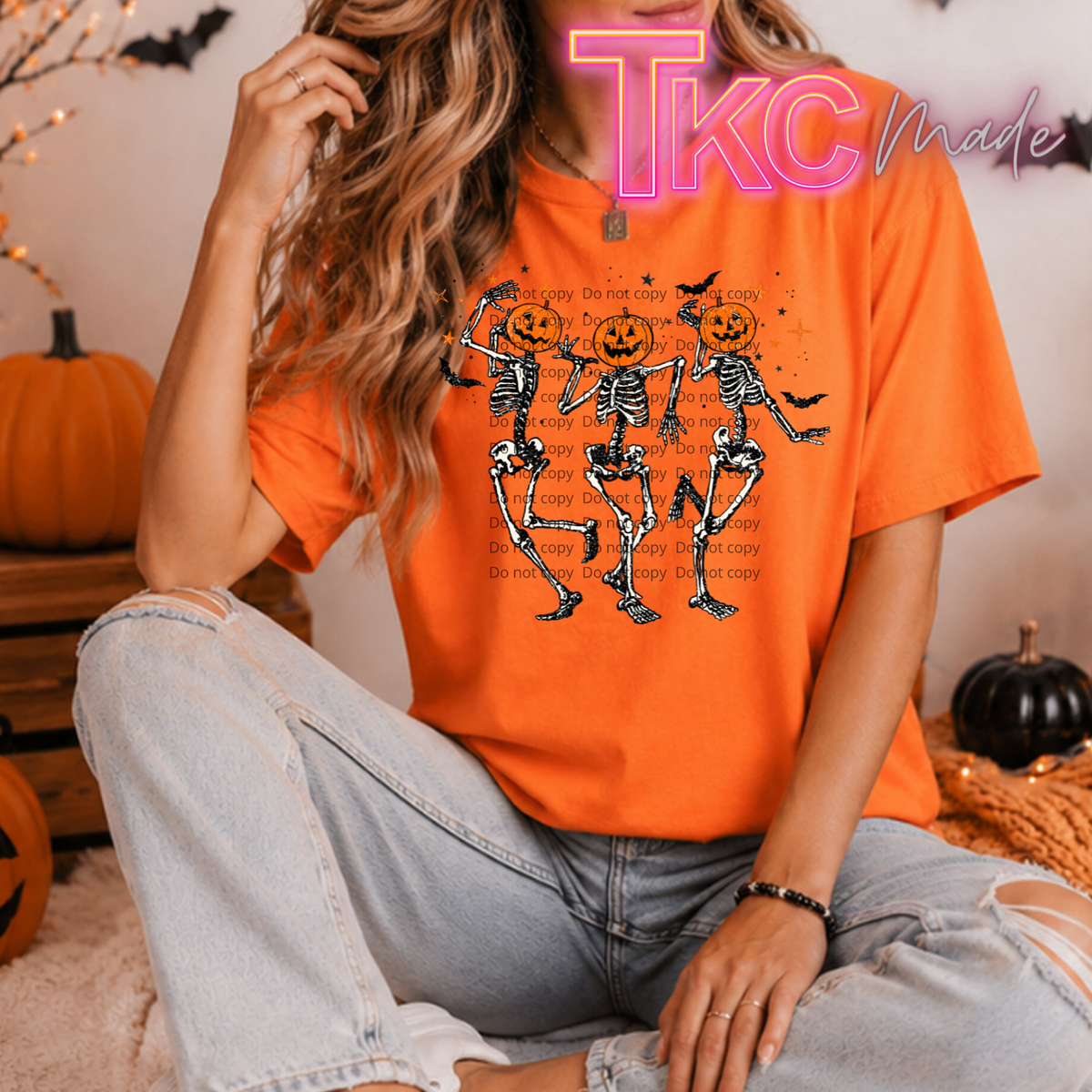 A woman wearing a bright orange graphic t-shirt with a Halloween design of three dancing skeleton pumpkins, paired with light-wash ripped jeans. The background is a cozy, decorated room with pumpkins and a 'TkC Made' watermark overlay.