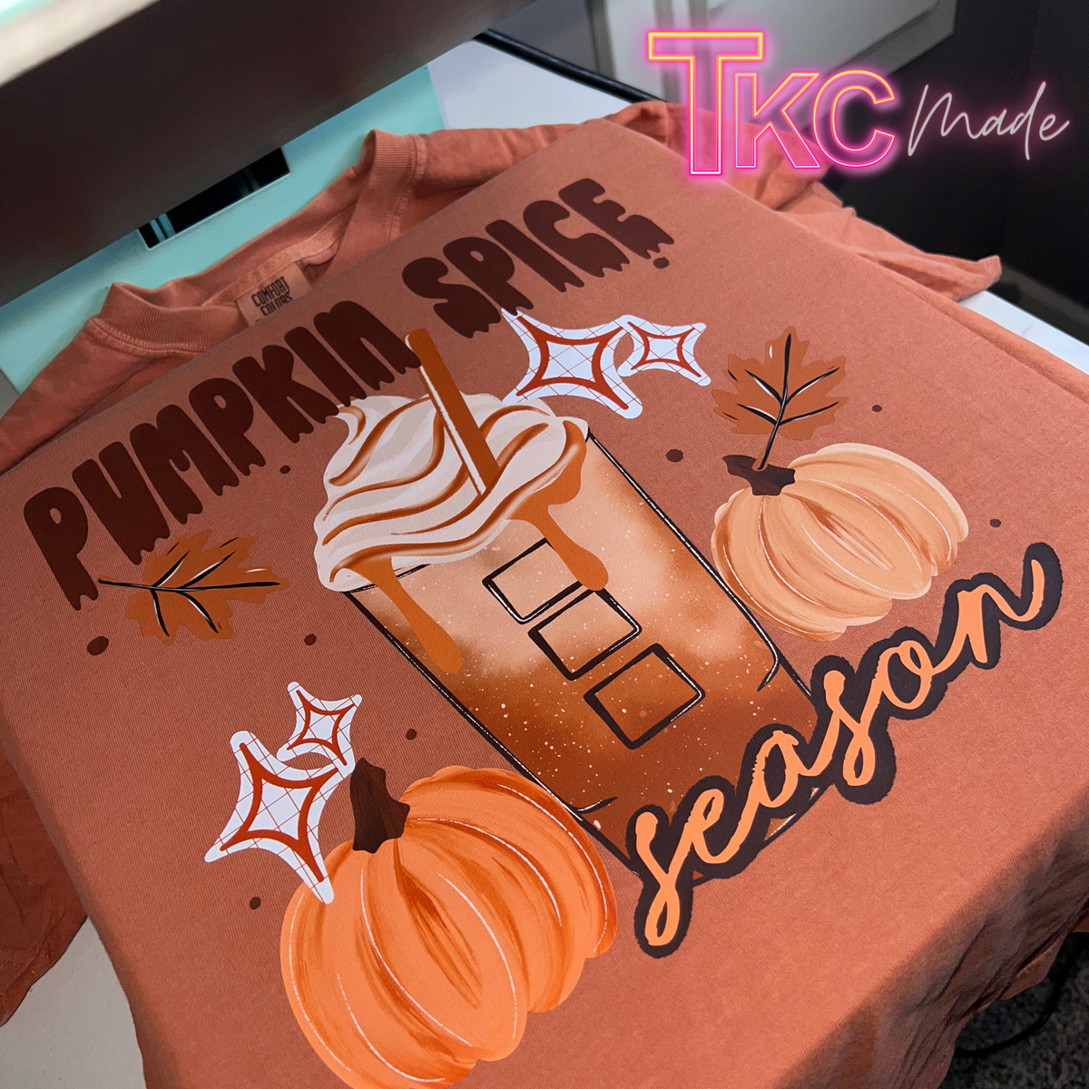 A rust-orange crewneck t-shirt featuring a 'Pumpkin Spice Season' graphic print with a pumpkin latte, pumpkins, and leaves, displayed flat on a light surface with a 'TKC Made' watermark in the corner.