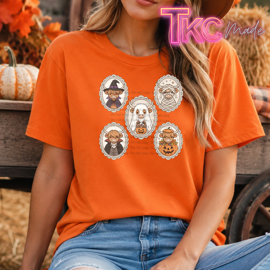 A woman wearing an orange graphic t-shirt with highland cows illustrations in Halloween outfits, set against a rustic fall-themed background with hay bales and a pumpkin.