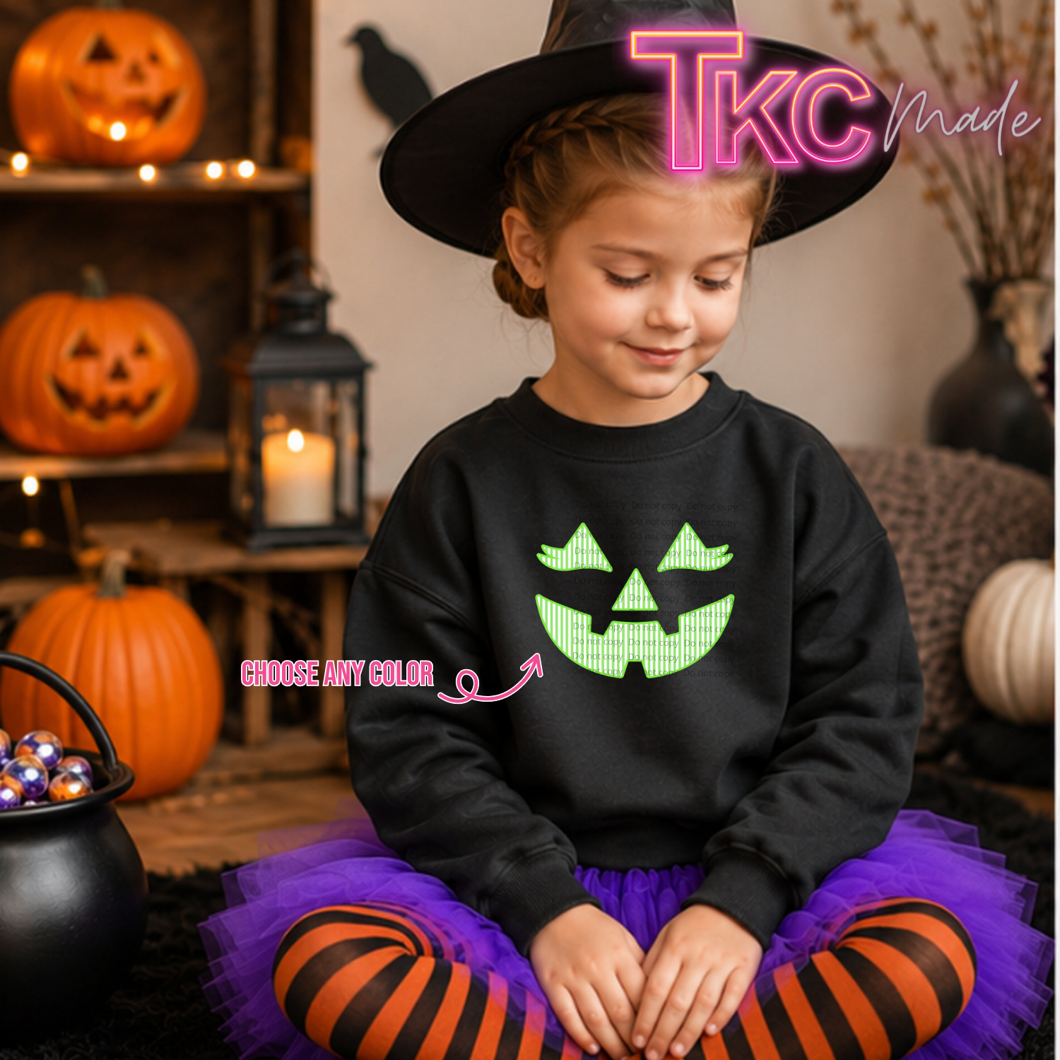 A young girl wearing a black Halloween-themed sweatshirt with a green jack-o'-lantern face graphic, a black witch hat, and a purple tutu with orange-and-black striped tights, sitting in a decorated Halloween room with pumpkins and a cauldron.