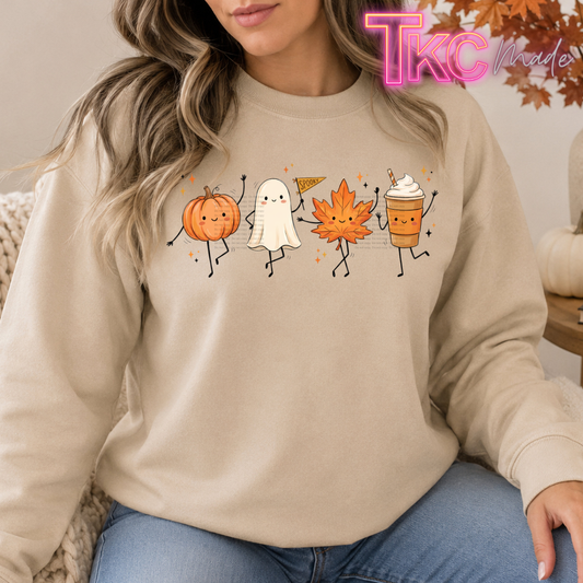A woman wearing a beige crewneck sweatshirt with a colorful autumn-themed graphic of a pumpkin, ghost, maple leaf, and coffee cup. The photo is taken in a cozy indoor setting with a 'TKC Made' watermark in the top right corner.