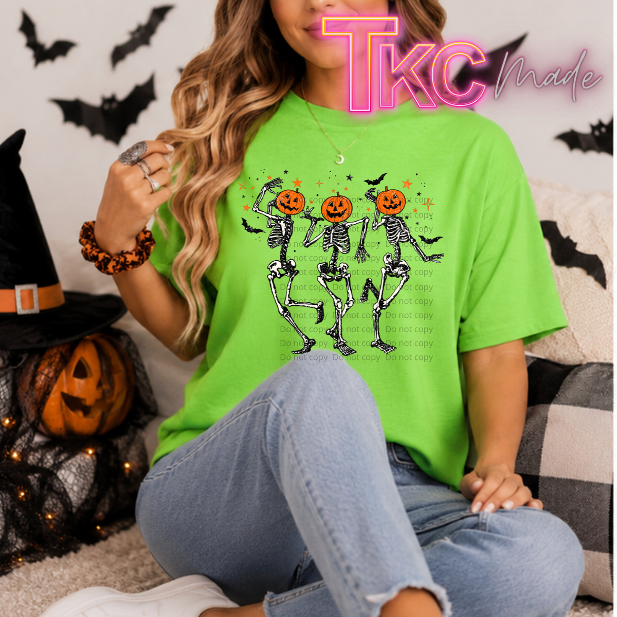 A woman wearing a bright lime green t-shirt with a graphic of three dancing skeletons holding orange jack-o'-lanterns, sitting on a couch with Halloween decorations.