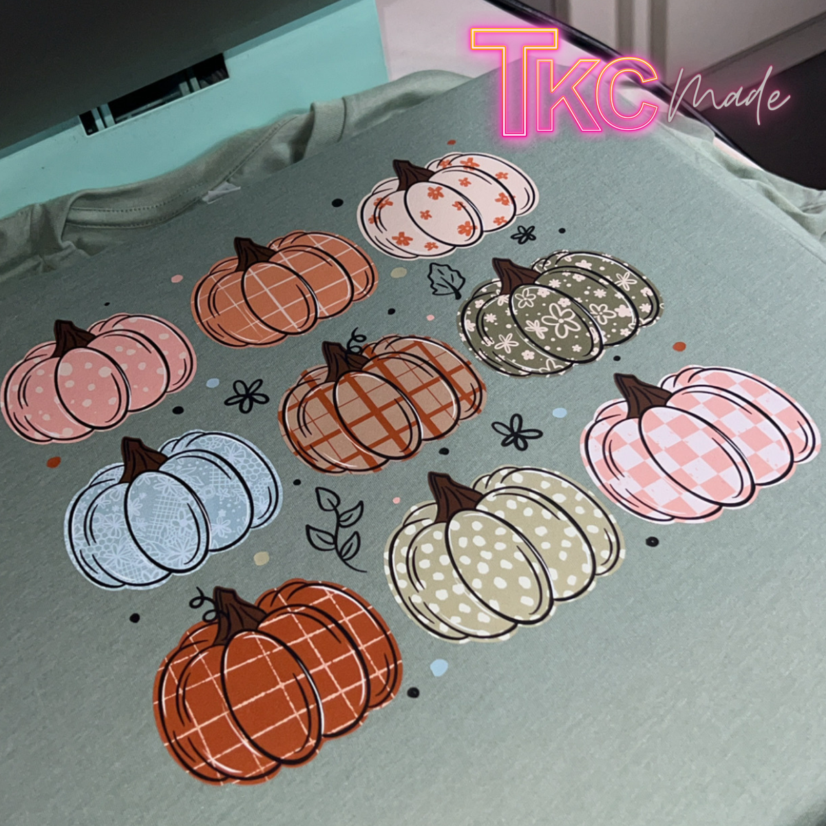 A close-up of a light sage green fabric printed with a pattern of nine autumn pumpkins in various colors and textures, including plaid, polka dots, and checkerboard patterns. A stylized 'TKC made' logo is visible in the top right corner.