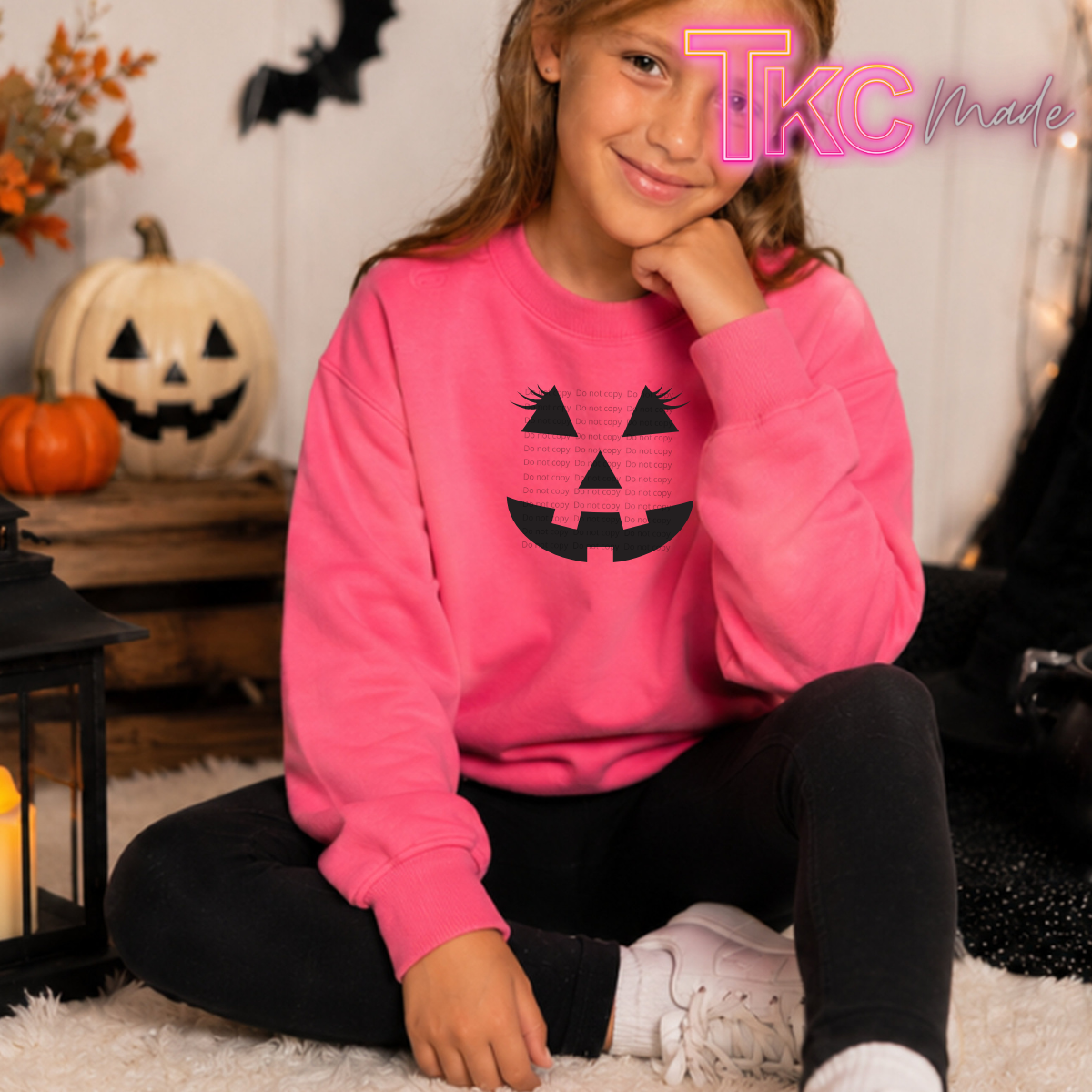 A young girl wearing a bright pink crewneck sweatshirt with a black jack-o'-lantern face graphic, sitting on a white rug in a cozy Halloween-themed room with pumpkins and a lantern.