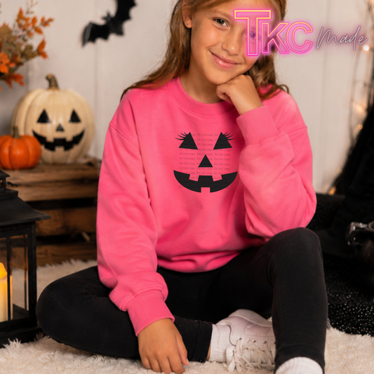 A young girl wearing a bright pink crewneck sweatshirt with a black jack-o'-lantern face graphic, sitting on a white rug in a cozy Halloween-themed room with pumpkins and a lantern.