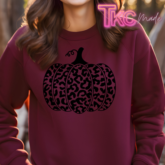 A person wearing a burgundy crewneck sweatshirt with a black leopard-print pumpkin graphic on the chest. The photo is taken outdoors with warm, backlit sunlight, and a pink 'TKC made' watermark is visible in the top right corner.
