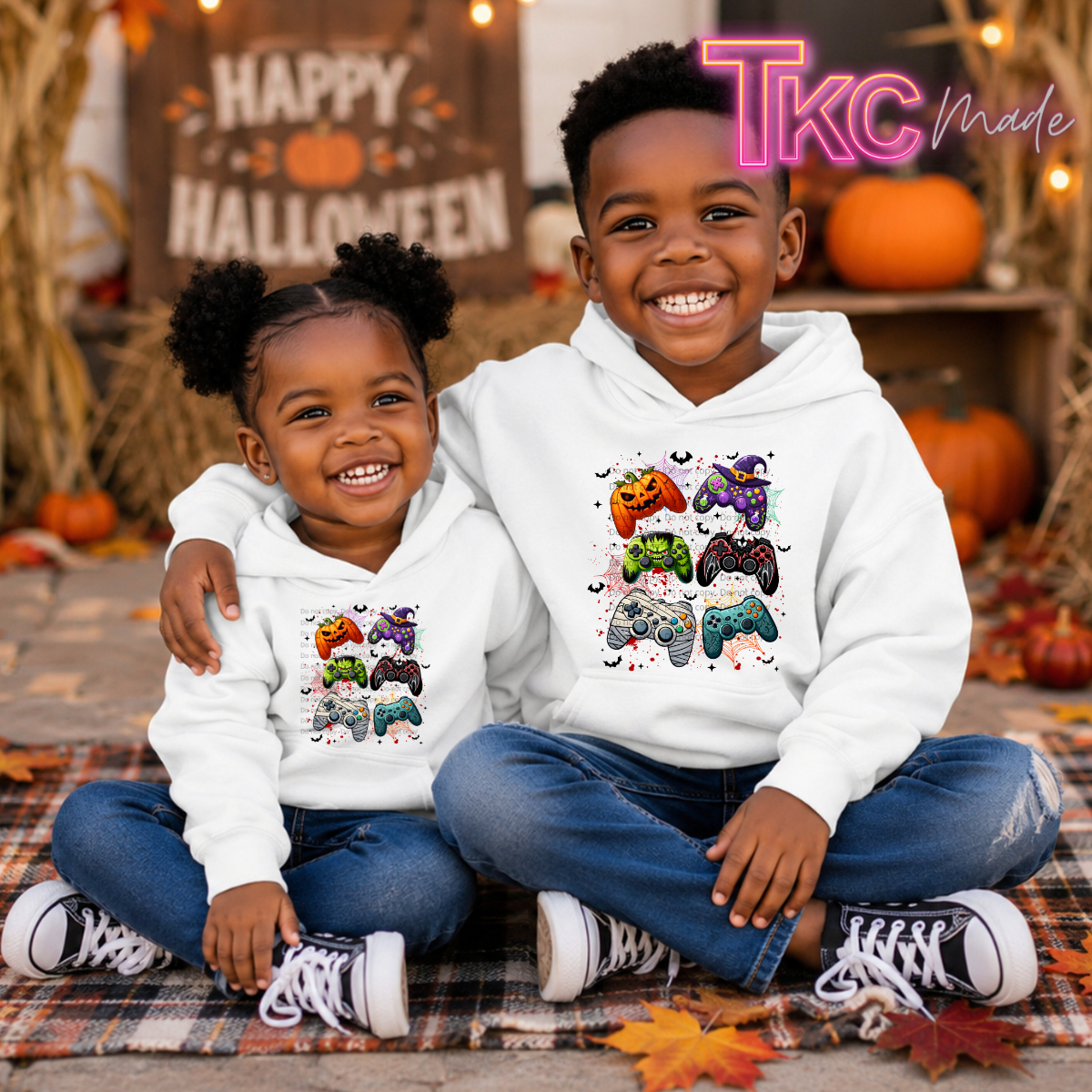Two children sitting together on a plaid blanket in a Halloween-themed setting, wearing matching white hoodies with colorful game controller graphics. The child on the right has a larger, more colorful graphic than the child on the left.
