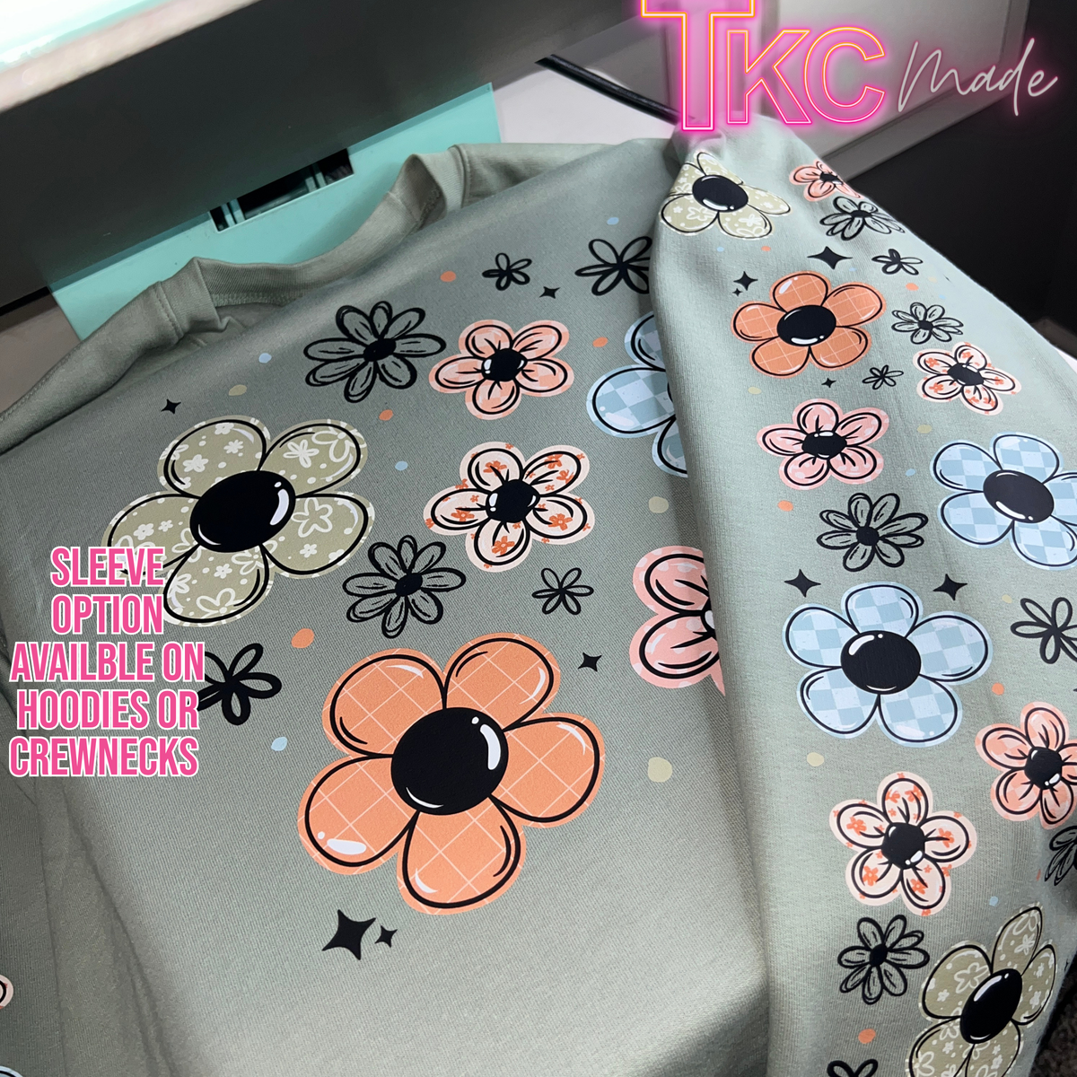 A sage green sweatshirt with a colorful floral pattern, laid flat on a desk. The pattern features large and small flowers in orange, light blue, pink, and beige with black outlines.