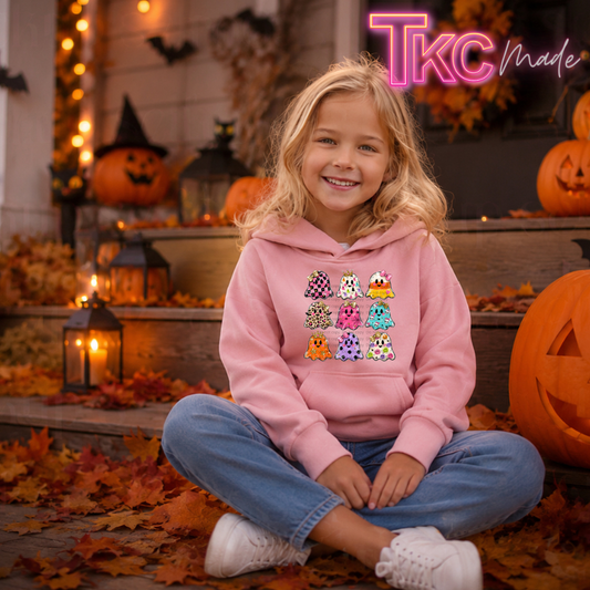 A young girl with blonde hair sits cross-legged on a porch covered in autumn leaves, wearing a pink hoodie with a colorful cartoon ghosts graphic on the front. The background features Halloween decorations including jack-o'-lanterns, lanterns, and warm string lights.