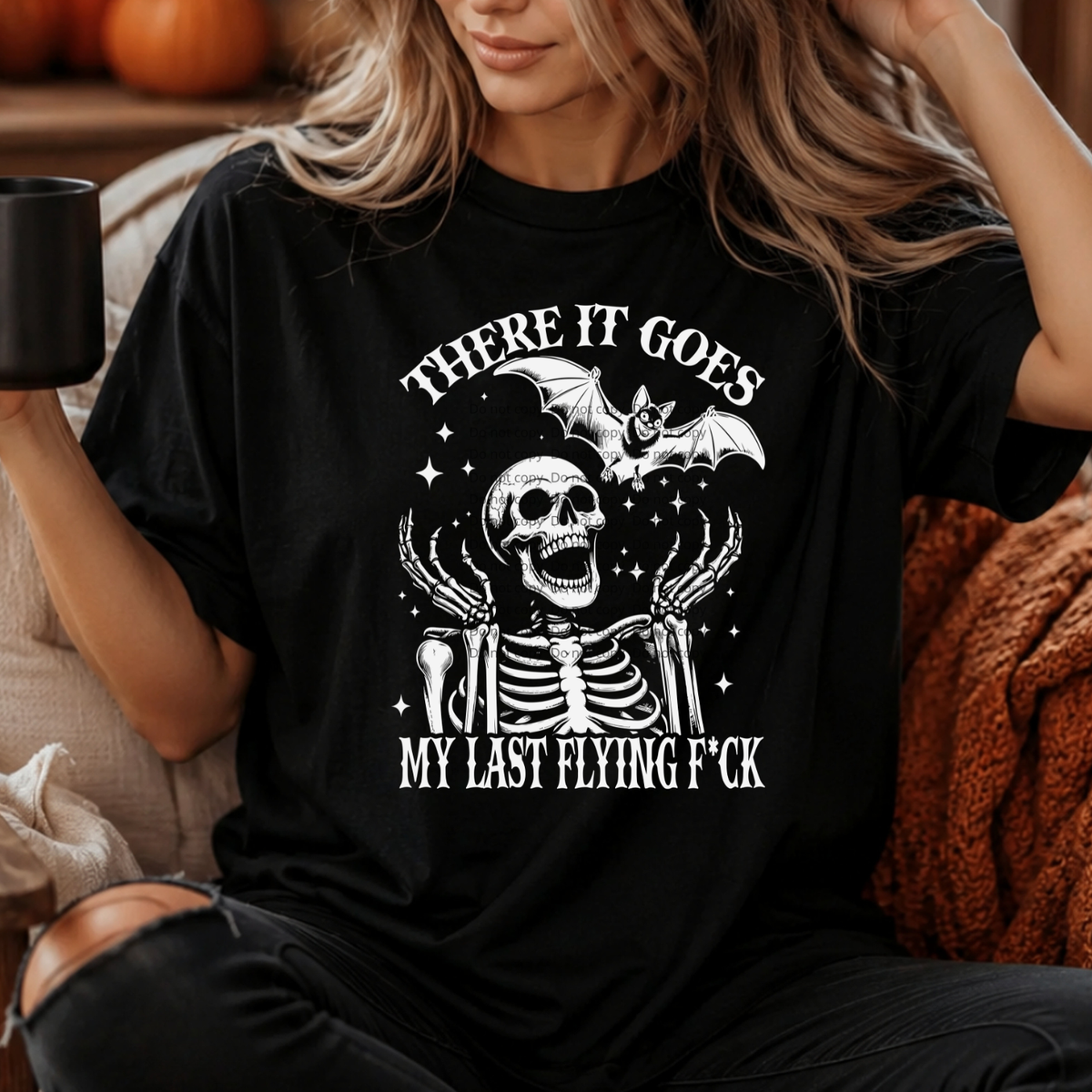 A woman wearing a black graphic t-shirt with a white skeleton and bat design, holding a black mug in a cozy indoor setting.