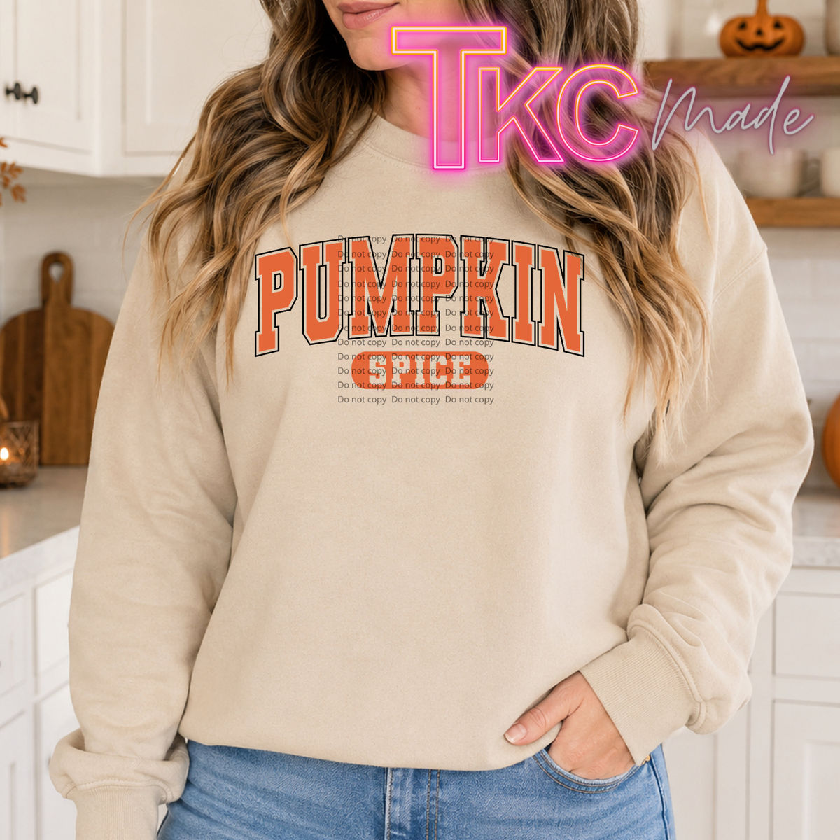 A woman wearing a cream-colored sweatshirt with 'PUMPKIN SPICE' printed in orange and brown block letters on the front, paired with blue jeans. The image has a 'TKC Made' watermark overlay.