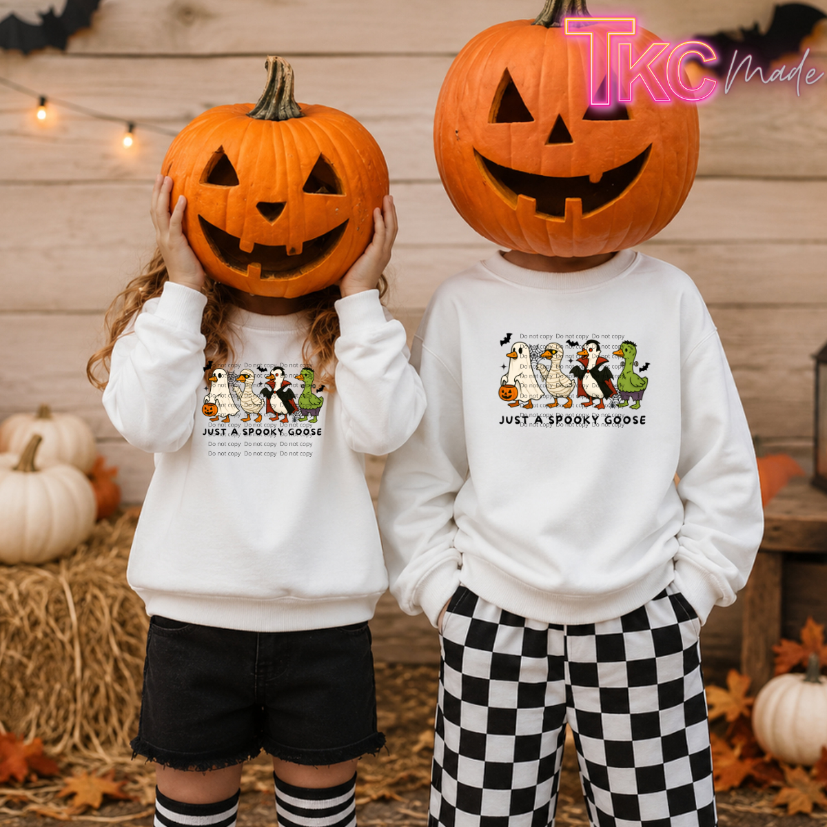Two children standing in a fall-themed setting wearing matching white sweatshirts with a cartoon spooktacular goose graphic and the text 'JUST A SPOOKY GOOSE'. The child on the left wears black shorts and striped socks, and the child on the right wears black-and-white checkered pants.