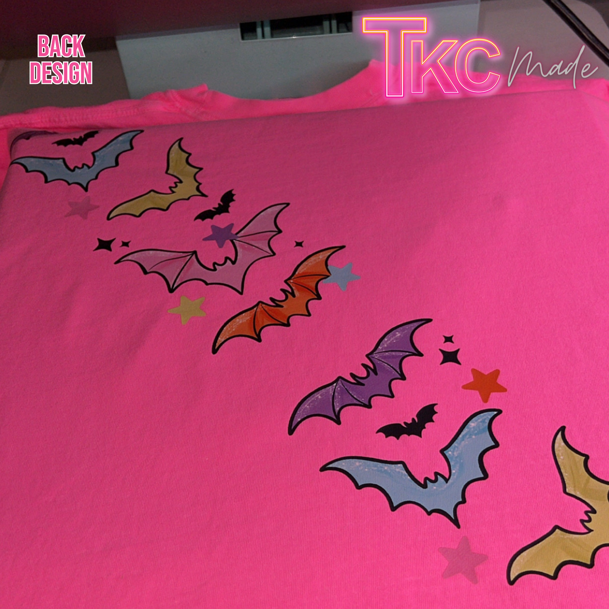 A bright pink t-shirt featuring a back graphic print of colorful bats and stars in a diagonal line. The image has 'TKC Made' and 'BACK DESIGN' text overlays.