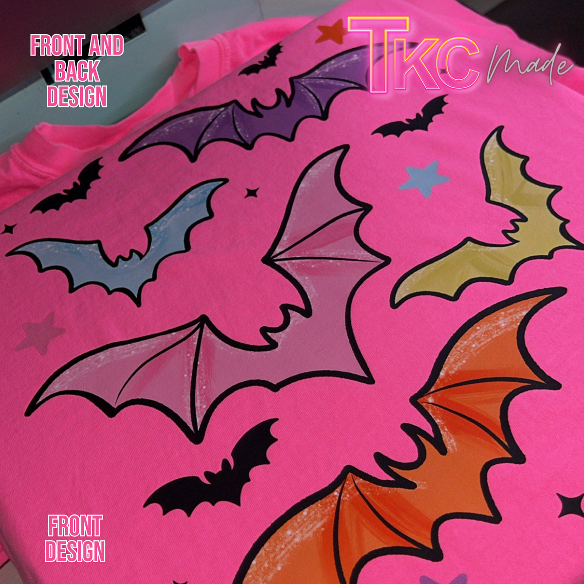 A close-up of a bright pink T-shirt with a Halloween-themed bat print. The design features multiple stylized bats in black, purple, blue, yellow, and orange, along with small black stars.