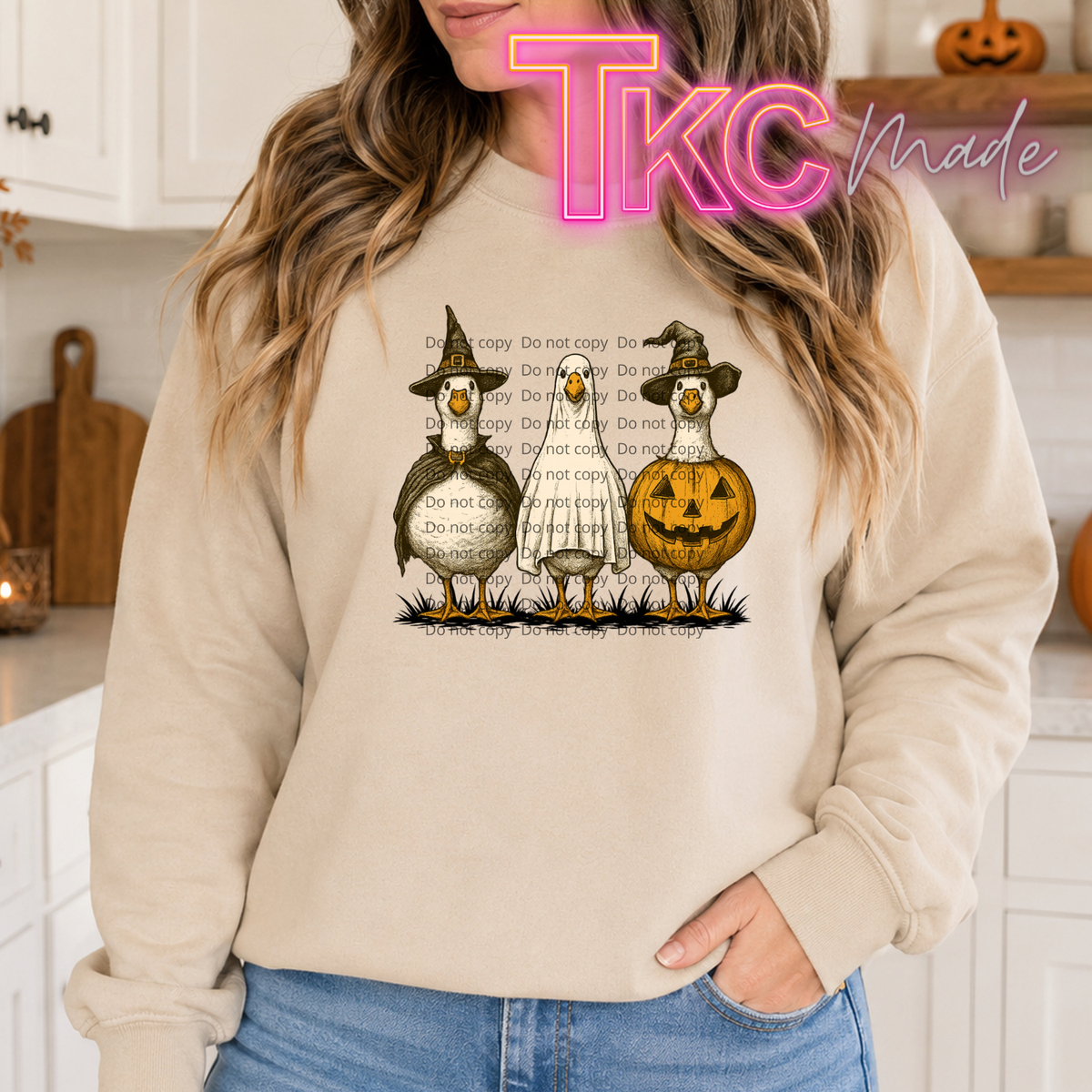 A woman wearing a beige sweatshirt with a Halloween graphic of three geese and a jack-o'-lantern, with a pink 'TKC Made' watermark overlay.