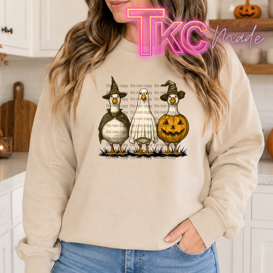 A woman wearing a beige sweatshirt with a Halloween graphic of three geese and a jack-o'-lantern, with a pink 'TKC Made' watermark overlay.