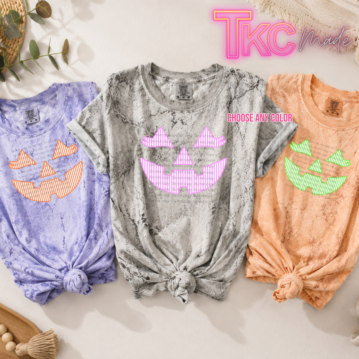 Three short-sleeve graphic T-shirts laid flat on a light beige surface, each with a knotted hem and a jack-o'-lantern face design. The center shirt is comfort color color blast with a purple jack O lantern, the left shirt is lavender with an orange jack o lantern, and the right shirt is orange with a green jack o lantern.