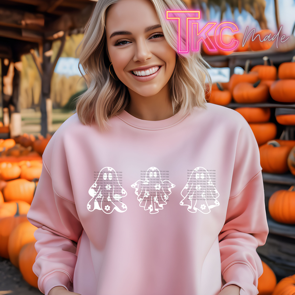A smiling blonde woman wearing a light pink sweatshirt with three white ghost graphics across the chest, standing in front of a pumpkin patch. A pink 'TKC Made' logo watermark is visible in the top right corner.