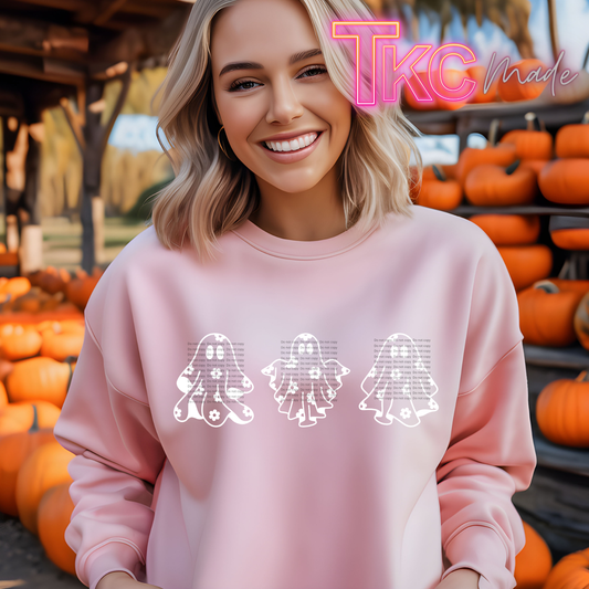 A smiling blonde woman wearing a light pink sweatshirt with three white ghost graphics across the chest, standing in front of a pumpkin patch. A pink 'TKC Made' logo watermark is visible in the top right corner.