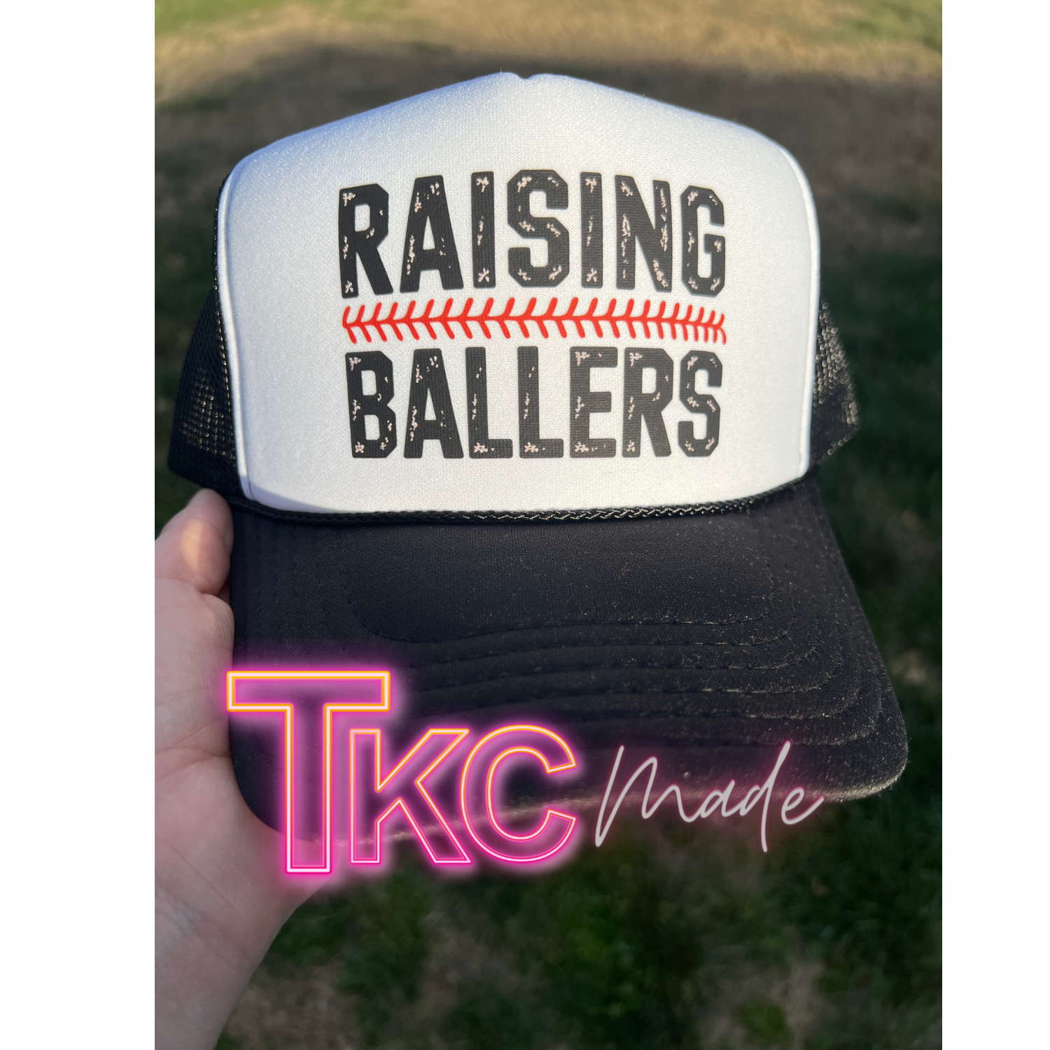 Trucker hat with 'RAISING BALLERS' text held by a hand, with 'TkC Made' branding.