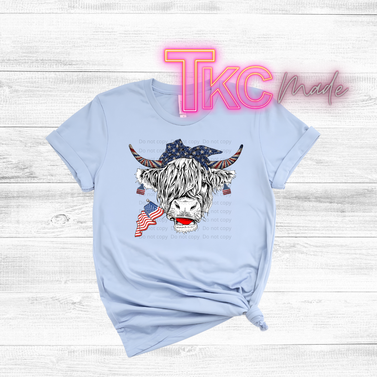 Light blue t-shirt with a graphic of a highland cow and American flag, on a wooden surface with 'TKC Made' text.