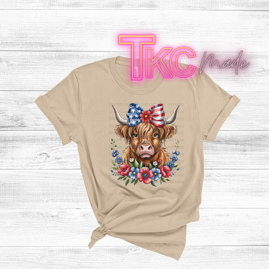 Beige t-shirt with a cow wearing a patriotic hat and flowers on a wooden surface with 'TKC Made' branding.