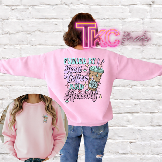 Pink sweatshirt with text fueled by iced coffee and anxiety the back,  and a iced coffee drink on the front pocket area. worn by a person against a white brick wall.