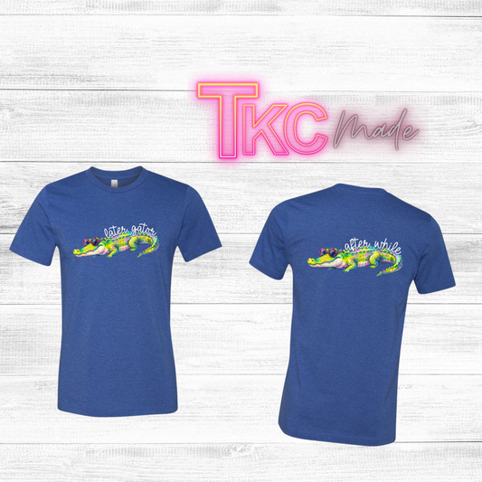 Blue t-shirt with a colorful gator graphic and text  that says later gator on the front and after while on the back. on a white wooden background with 'TKC Made' branding.