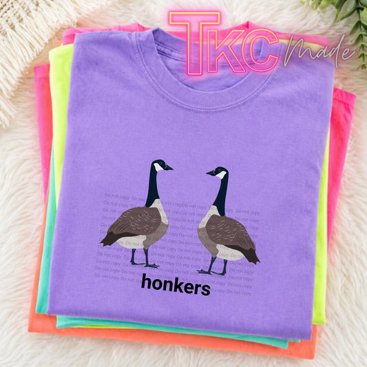 Stack of colorful t-shirts with geese design and 'honkers' text on a textured surface.