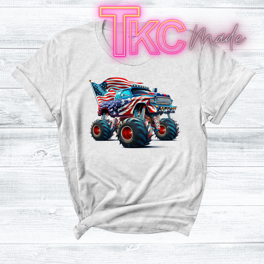 White t-shirt with a graphic of a monster truck with an American flag design, on a light wooden surface with 'TKC made' text.