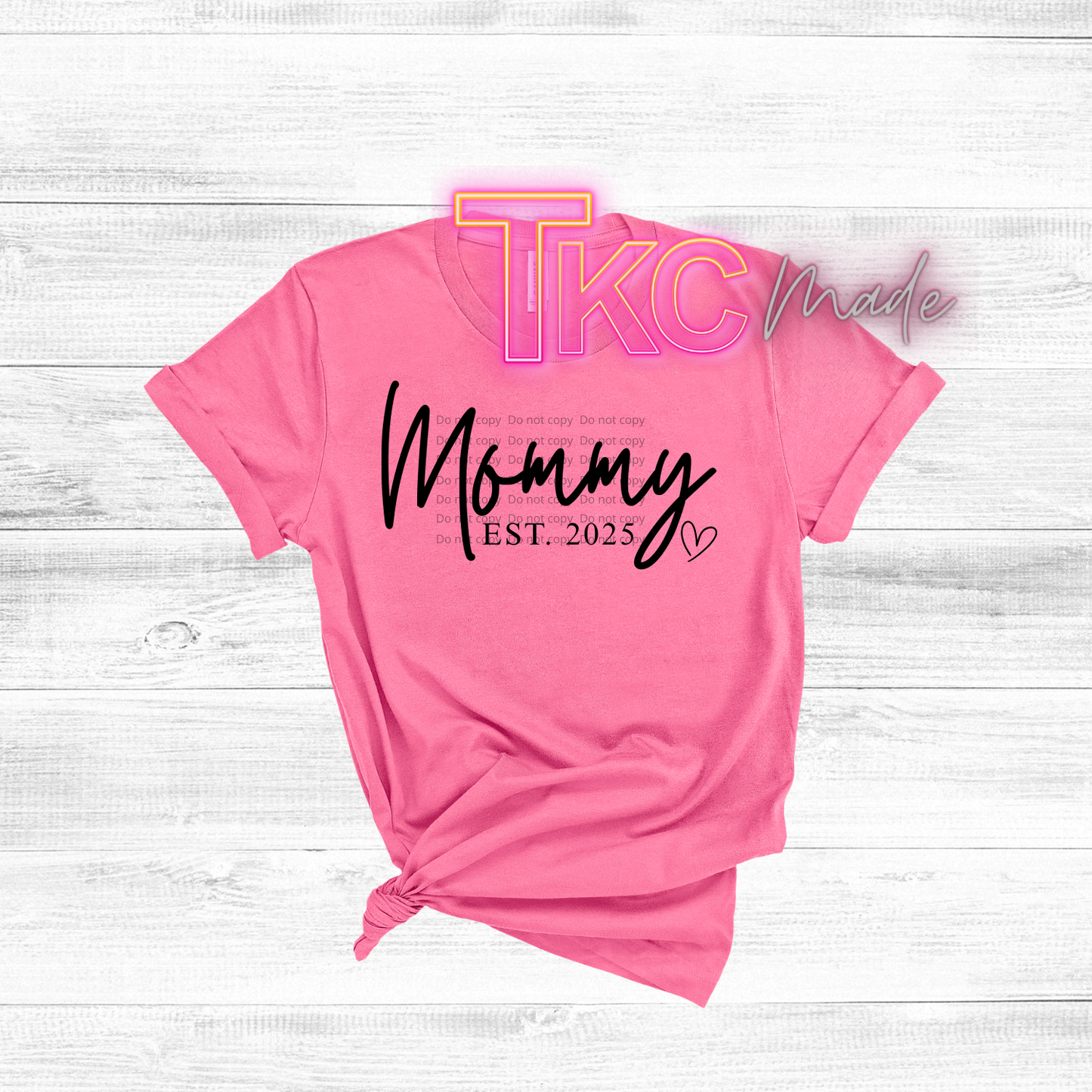 Pink t-shirt with 'Mommy EST. 2025' text on a white wooden surface