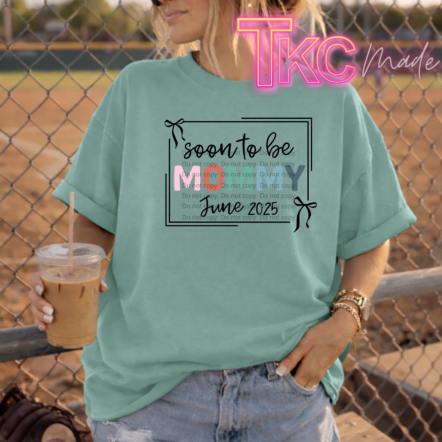 Person wearing a green t-shirt with a pregnancy announcement design, holding a drink, with a neon sign in the background.