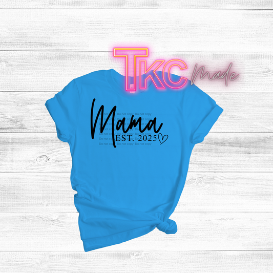 Blue t-shirt with 'Mama EST. 2025' text on a white wooden surface, featuring 'TKC made' branding.