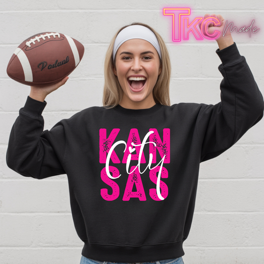 A woman wearing a black sweatshirt with a pink and white 'Kansas City' graphic stands against a white brick wall, holding a brown football in one hand and raising the other arm. A 'TKC Made' watermark is visible in the top right corner.