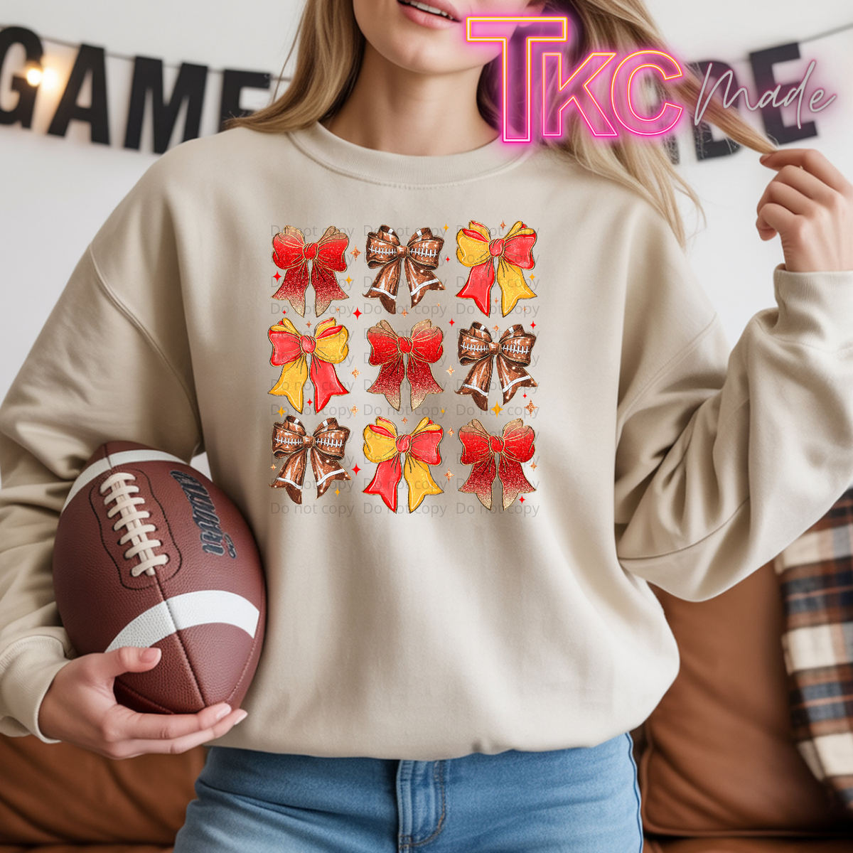 A woman wearing a beige sweatshirt with a graphic print of nine bows in red,  yellow, and footballs. She is holding a brown football in her hand, with a couch and a 'TKC Made' watermark in the background.