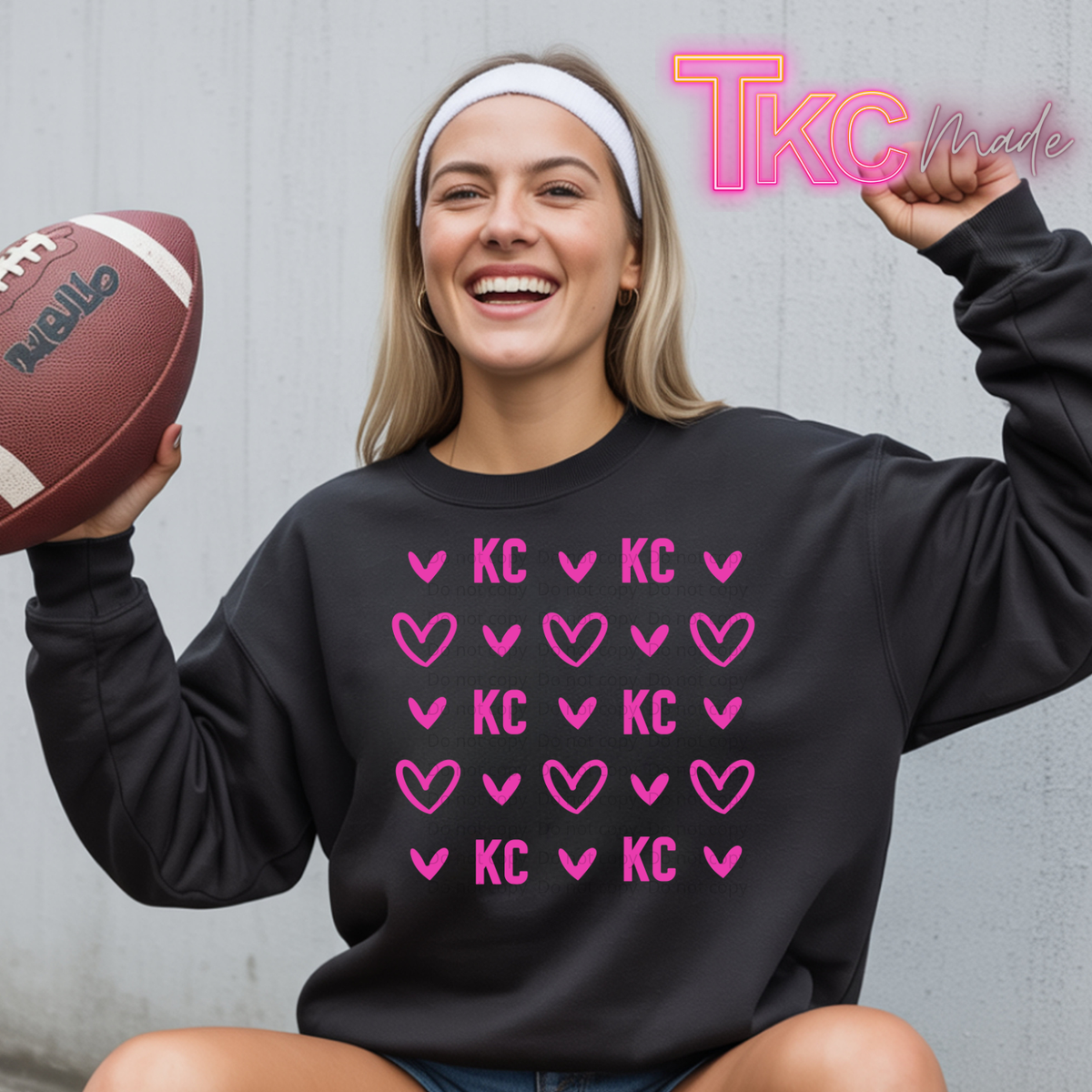 A woman wearing a black sweatshirt with pink heart and KC lettering, holding a football and posing against a gray wall. A 'TKC Made' logo is visible in the top right corner.