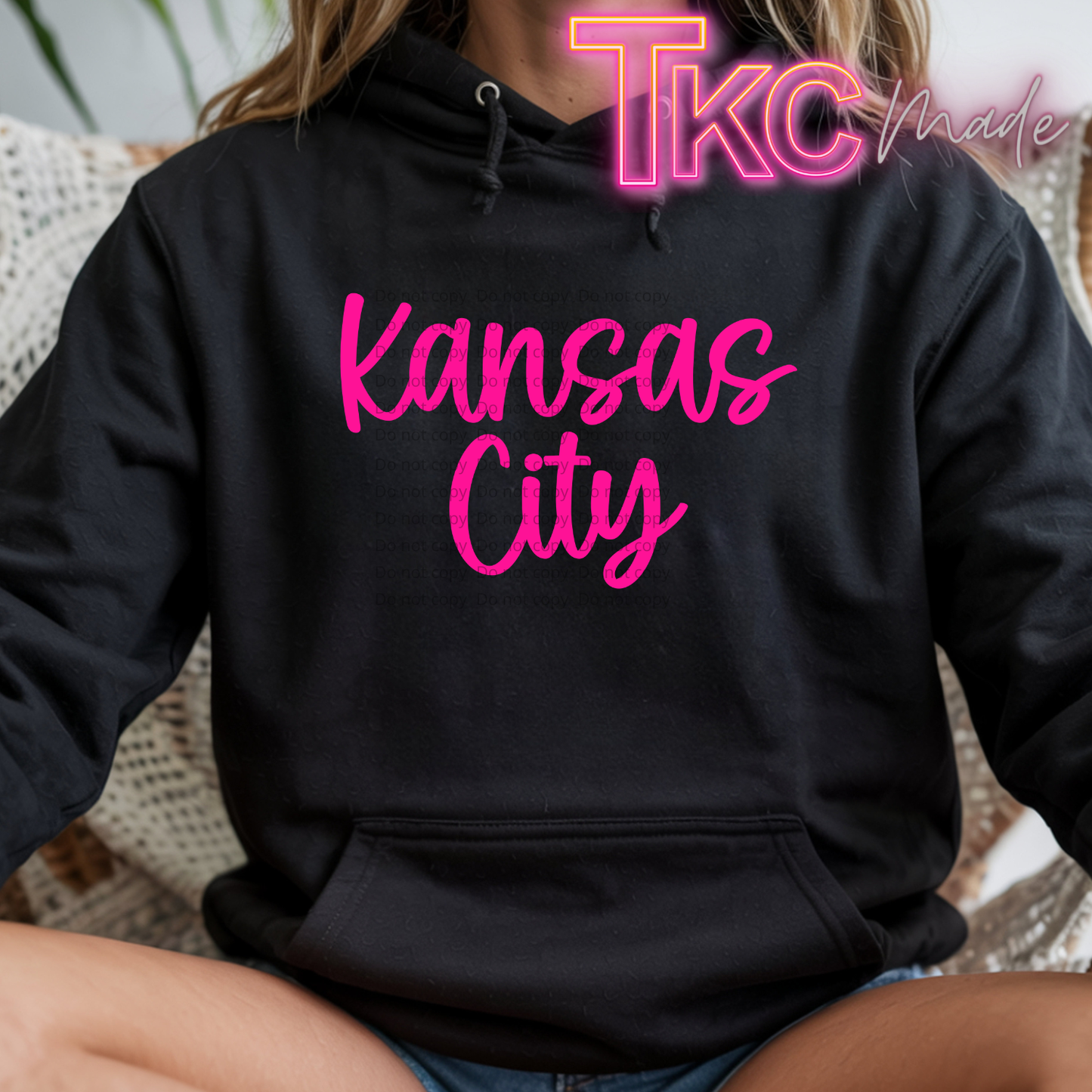 A person wearing a black pullover hoodie with 'Kansas City' printed in bright pink script across the chest. The photo is cropped from the neck down, showing the person sitting indoors on a woven chair.