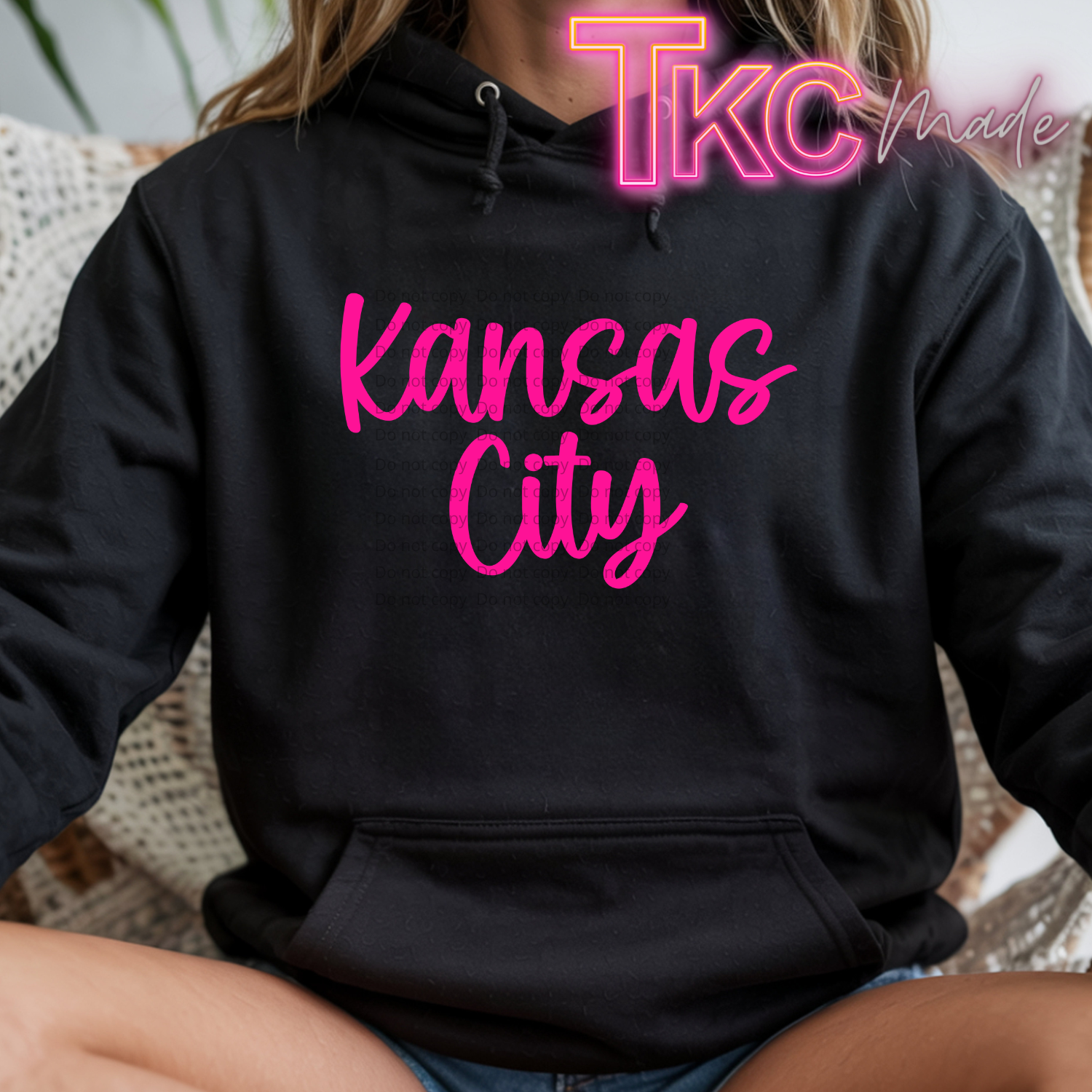 A person wearing a black pullover hoodie with 'Kansas City' printed in bright pink script across the chest. The photo is cropped from the neck down, showing the person sitting indoors on a woven chair.