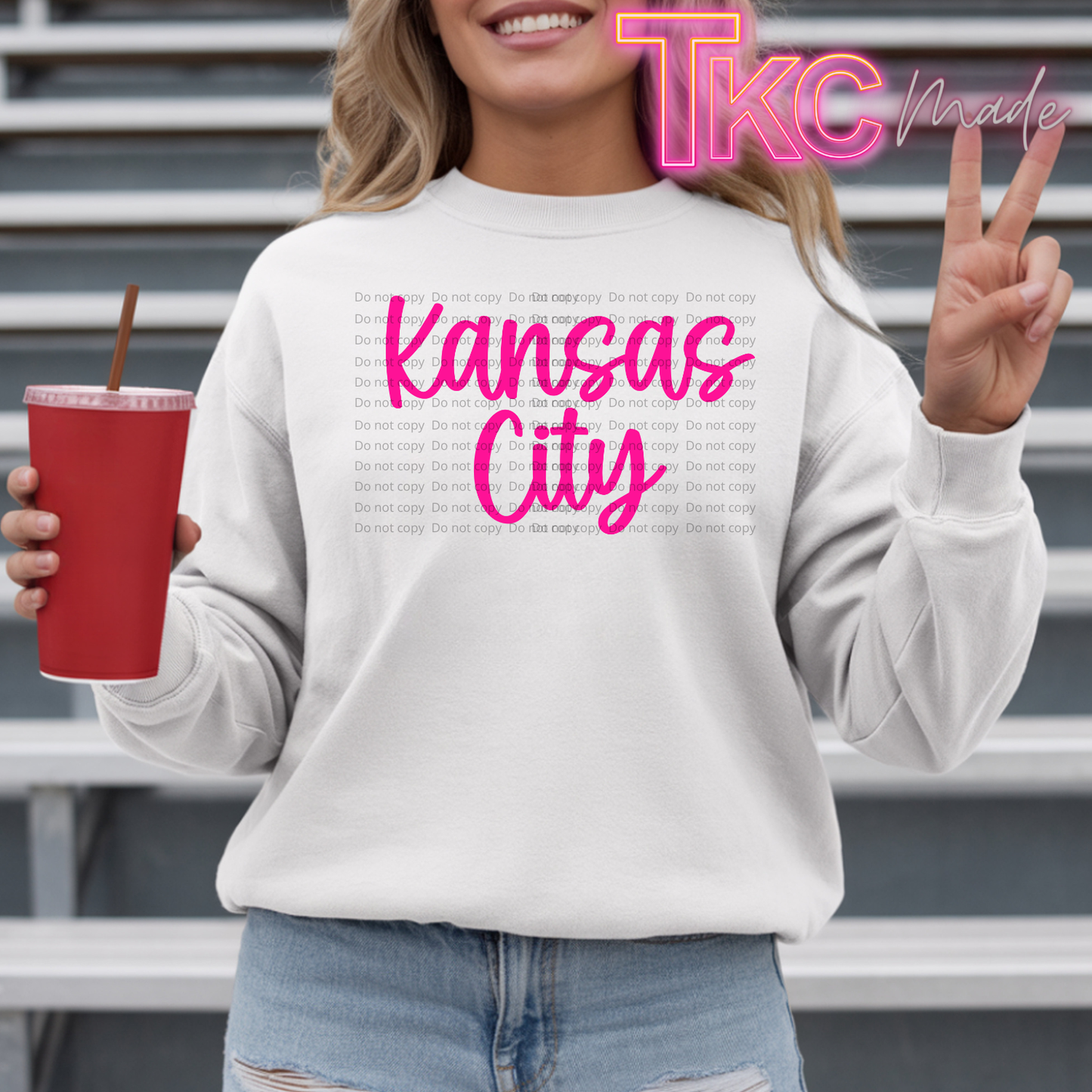 A woman wearing a white sweatshirt with a pink 'Kansas City' graphic and a faint city skyline print, holding a red cup in front of gray metal steps. A 'TKC Made' watermark is visible.