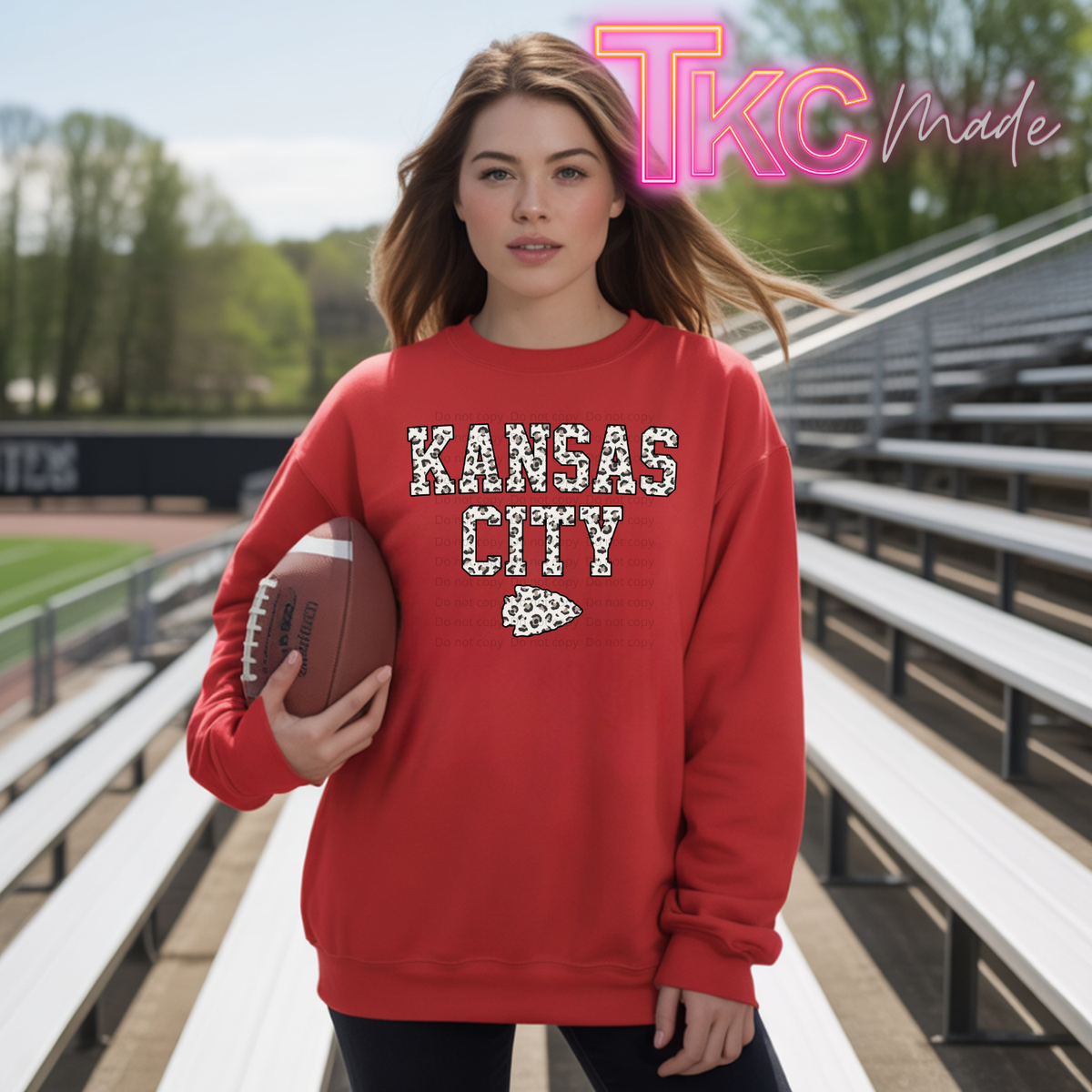 A woman stands on outdoor stadium bleachers holding a football, wearing a red sweatshirt with 'KANSAS CITY' and the Kansas City Chiefs logo in white and black patterned lettering. A pink 'TKC Made' watermark is visible in the top right.